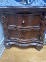 Close-up of front view of wooden night table showing two drawers with curved fronts and round metal knobs, carved moldings and stylized legs