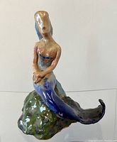 Front angled view showing mermaid sitting on textured green rock with blue/black tail and beige upper body.