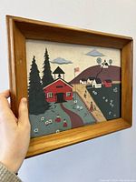 Framed folk art painting showing red schoolhouse, pine trees, and rural homes on hills with path and figures.