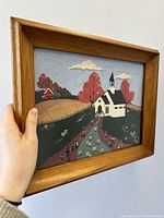 Hand holding the white oak framed folk art painting showing the church, red trees, and pathway with figures.
