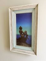 Framed vertical art print showing the mermaid figure on a rock against a blue starry sky, set in a textured pale mat and rustic white frame with slight wear.