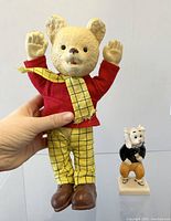 Plush Rupert Bear doll held upright with hand, showing red top, yellow checkered pants, scarf, and brown boots, with visible wear on pants.