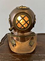 Front view of bronzed metal diver's helmet desk lamp with lit bulb visible through crosshatched window.