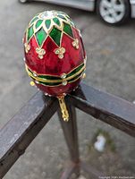 Close-up view of the red and green egg-shaped musical ring box with jeweled floral accents and tri-footed gold-colored stand, showing its ornate and glossy finish.