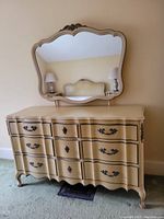 Off-white painted wood dresser with serpentine front and ornate black metal handles, topped by matching scalloped mirror with carved crest.