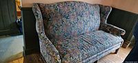 Floral patterned high back 3-seater couch with padded arms and three seat cushions in a room corner.