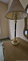 Full view of brass base table lamp with black column and beige lampshade placed on a beige brown surface indoors near a window and mirror.