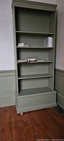 Full front view of the tall sage green wooden bookcase with shelves and base drawer.