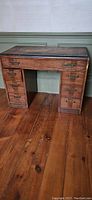 Photo shows a vintage wooden desk with six drawers on the sides and one center drawer, placed on a wooden floor against a wall.