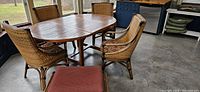 Oval wooden table with slatted top surrounded by four woven wicker chairs with padded seat cushions
