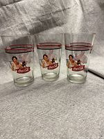 Three clear Coca Cola Gibson Girl vintage tumbler glasses with red, white, and black stripe decoration and classic Gibson Girl illustration.