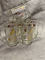 Six frosted glass beer mugs featuring golden etched pine cone and acorn designs with gold rim detailing arranged on fabric.