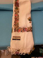 Folded off-white pashmina shawl showing colorful embroidered floral border and label tag reading 'Kashmiri Pashmina Shawls Dry Clean Only'.