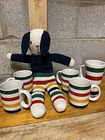 Set of 4 Hudson Bay coffee mugs and a knitted stuffed dog toy wearing Hudson Bay striped pants and navy sweater, displayed on a wooden plank against a gray concrete block wall.