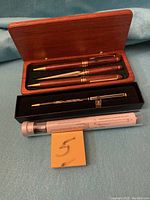 Photo showing five vintage pens including two wood-grain finish pens in wooden case, Ford pen in black box, pen in plastic tube, and Lenze mechanical pencil.