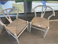 Two white Wishbone style chairs with woven jute seats placed indoors near glass windows. The chairs have white painted wooden frames and natural colored woven jute seats. Visible base wear from cat damage repaired by sanding and repainting.