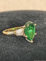 Close-up of pear-shaped faceted green stone set in gold plated silver ring with baguette side stones on black background.