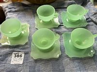 Five vintage jadeite teacups arranged on their matching square saucers on fabric.