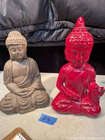 Photo of two Buddha statues side by side, one gray stone-colored, one red glossy, placed on a surface with dark background.