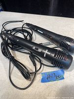 Two black Sylvania microphones with attached cables laid out flat. Both have a metal mesh grille, on/off switch, and 1/4 inch audio jacks.