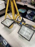 Close-up of two rectangular work lights with metal protective grids and electric cords, mounted on the yellow tripod stand.