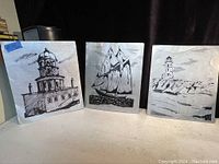 Three aluminum sheets with black printed images of a clock tower lighthouse, a sailing ship, and a lighthouse on a rocky shore