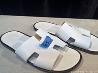 Left side angle view of the white slide sandals showing the cutout H-shaped pattern on the strap and the flat sole.