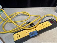 Photo of a black power strip with six outlets, yellow cord, on/off switch with red light, placed on a light-colored surface, showing the entire length of cord.