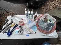 Photo of assorted tools and hardware on white table including caulking guns, sealants, mounting brackets, hoses, tape, and miscellaneous hardware items.