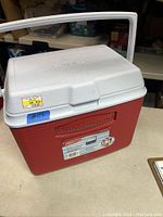 Side angle view of the entire cooler showing the red ribbed body, white hinged lid, carrying handle, and front label.