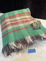 Folded vintage wool blanket showing green, brown, beige, and red tartan pattern with fringed edge