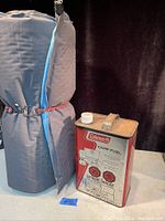 Therm-a-Rest sleeping pad rolled and secured next to a one-gallon Coleman camp fuel can.