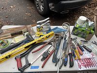 Photo showing various hand tools laid on a plastic table outdoors. Includes spirit level, hand saw, hammer, pipe wrench, pliers, staple gun, screwdrivers, wire brushes, measuring tape, and drill bits.