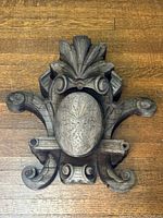 Top-down view showing full carved wood architectural shield with oval medallion and surrounding scroll and leaf details on wood floor.