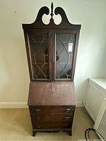 Front view of antique secretary desk showing decorative top finial, glass paneled doors with geometric woodwork, drop front panel, and three drawers below.