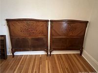 Pair of antique wooden single bed headboards side by side against wall showing full front view with parquet pattern and floral inlay.