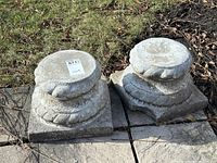 Two small square cement bases with rounded tops and rope-like decorative indentations, positioned outdoors on stone tiles surrounded by grass and dry leaves.