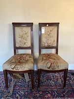 Front view of both antique wooden chairs with floral upholstery showing wear and tape on metal casting on one chair.