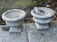 Pair of vintage concrete urn style planters side by side on stone surface outdoors.