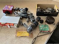 Photo showing red tester device, assorted radio tubes scattered, boxed tubes, Bulldog Elecsosnip packaging, and a round brown component with cable.
