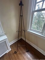 Full view of the vintage wrought iron art easel standing on a wooden floor next to a window and radiator, showing its tall tripod shape and ornate fleur-de-lis top.