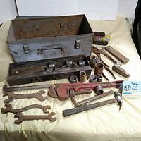 Metal toolbox and assorted hand tools laid out including pipe wrench, hammers, and wrenches