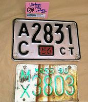 Top view of two metal license plates. The upper plate is a 1956 Connecticut plate, white with black lettering and a red vintage date sticker. The bottom plate is a Massachusetts 1990 plate with white background and green embossed numbers, showing some rust and discoloration.