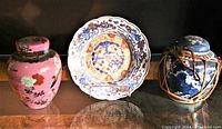 Lot of three items including pink ginger jar, blue ginger jar in woven casing, and decorative bowl plate with floral patterns.