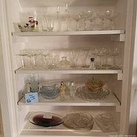 Lot of clear and colored glassware on white shelves including stemware, bowls, small plates, and decorative glasses.