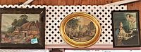Three paintings displayed on a lattice background, one oval golden frame village street scene in center, flanked by two rectangular wood framed paintings: scenic cottage on left, religious piano scene on right.