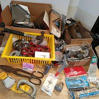Overview of assorted handyman tools and hardware items in boxes and a yellow basket including hammer, tin snips, locks, tape measure, and more.