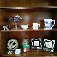 Shelf view showing all 10 vintage collectible items arranged with focus on glass compote, pitcher, bowls, framed prints, woven ball, and jar