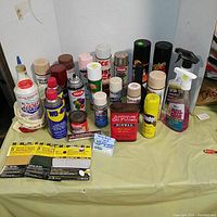 Overview of the lot showing 15 assorted car maintenance products including spray paints, lubricants, and sanding discs arranged on a table