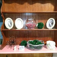 Photo showing the lot arranged on wooden shelving and pink surface including oval picture frames, glass vase, green round accents, black and gold spherical accent, silver basket with foliage, and ceramic cherub plant holder.